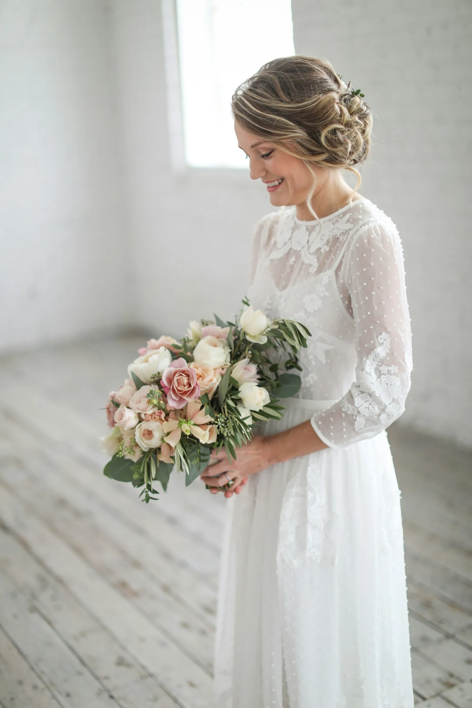 Noiva de perfil com vestido branco de renda e mangas, segurando um buquê delicado