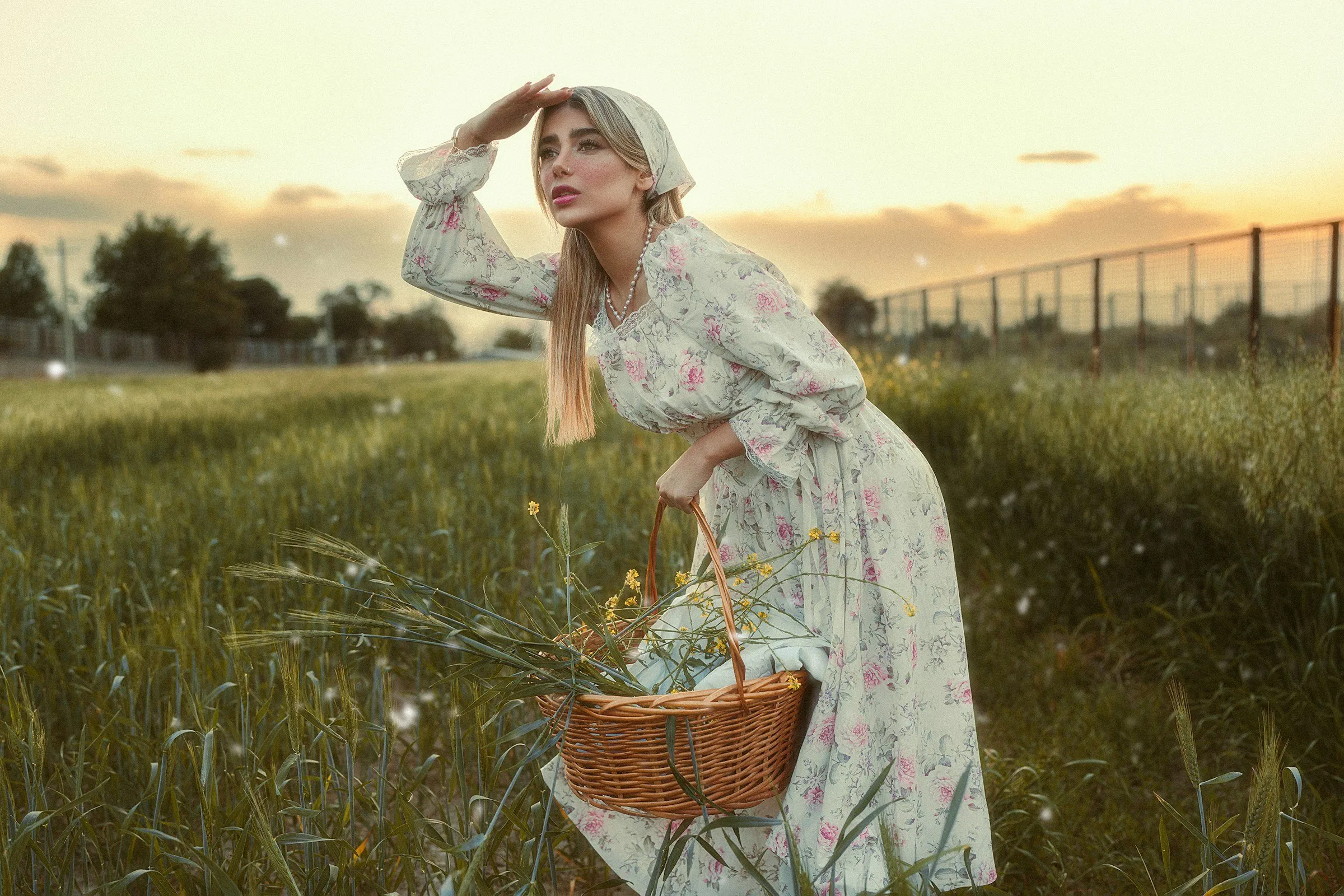 Mulher com vestido longo floral e lenço no cabelo em um campo ao pôr do sol, segurando uma cesta
